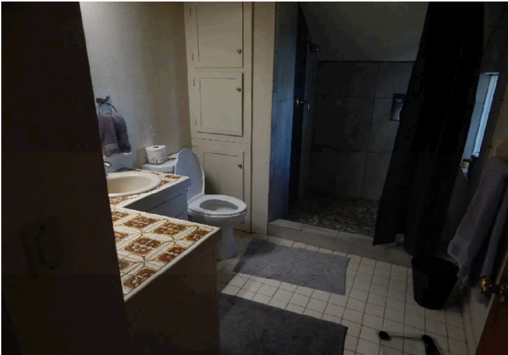 Bathroom before renovation with tile counters, dated cabinetry, and dark shower stall